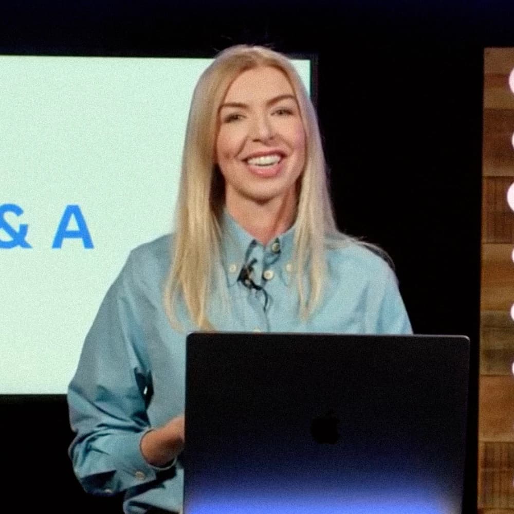 Tara Capella McLaughlin, a blonde woman in an oxford button down standing on stage with a large kaynote presentation behind her.